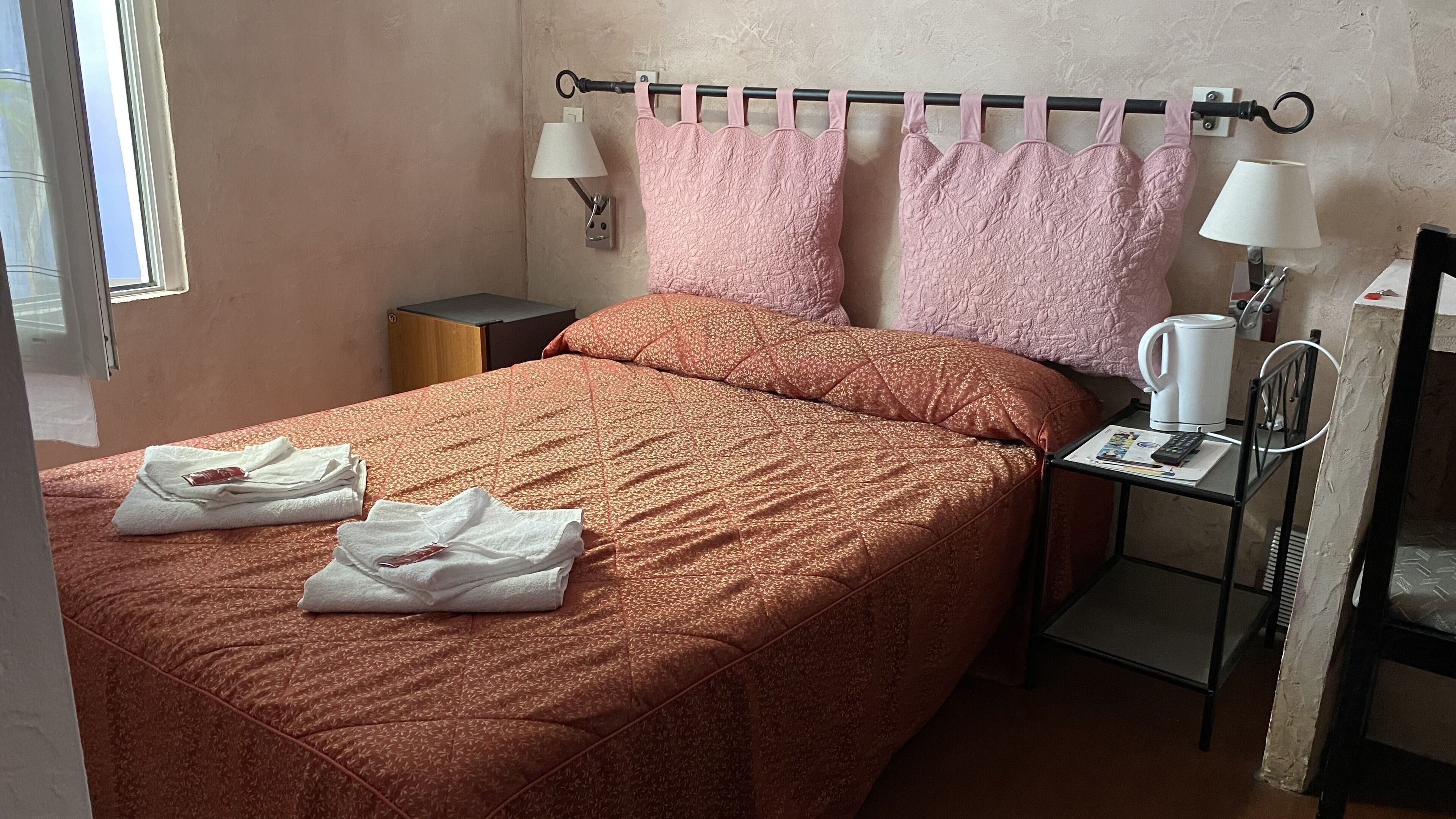 A neatly made bed with a patterned bedspread, two pillows, and folded towels on the bed. There is a nightstand with a lamp, a small table with a kettle, and a chair.
