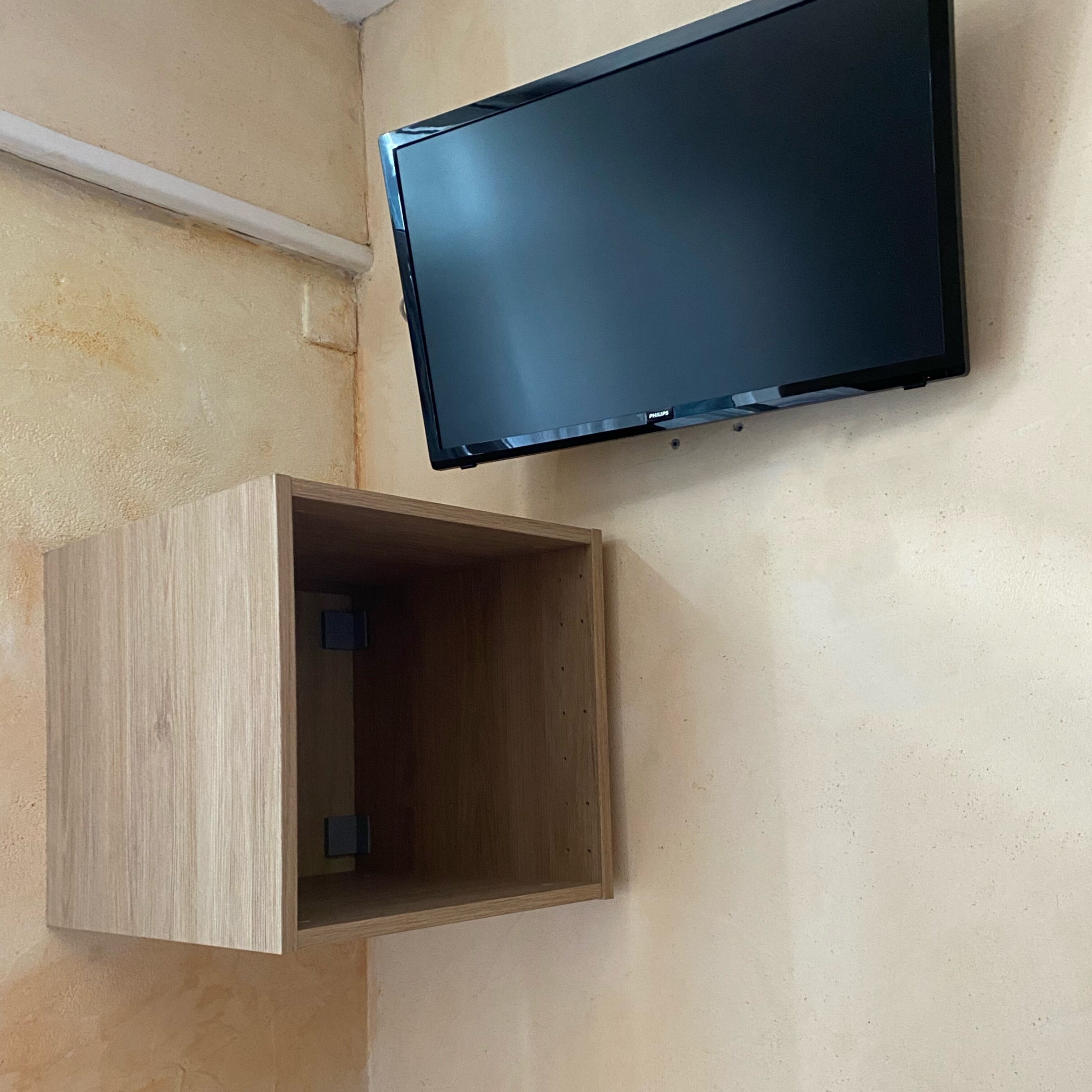 Uma televisão fixada na parede e um armário de madeira, tudo contra um fundo bege.