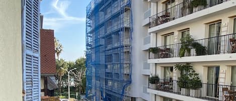 A view of a building under construction with scaffolding, a street with a parked car, and a balcony with a potted plant.