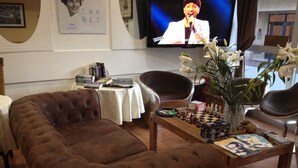 A cozy living room with a brown tufted sofa, a coffee table with chess pieces, a television displaying a show, and a vase with white flowers. - Boutique Hôtel Couleurs du Sud (Cannes)