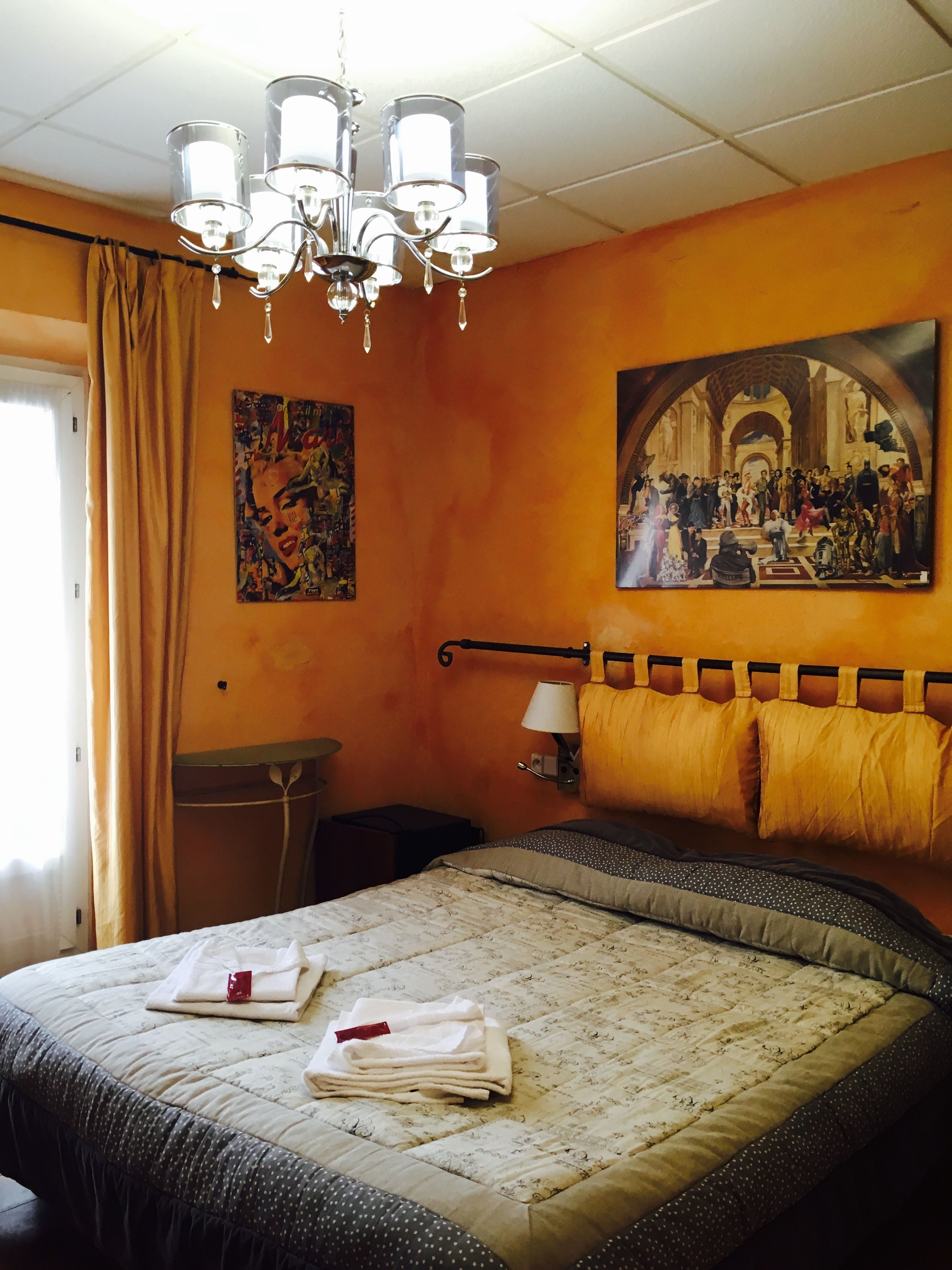 Une chambre d’hôtel avec un lit, un tableau, un lustre et une fenêtre avec des rideaux.