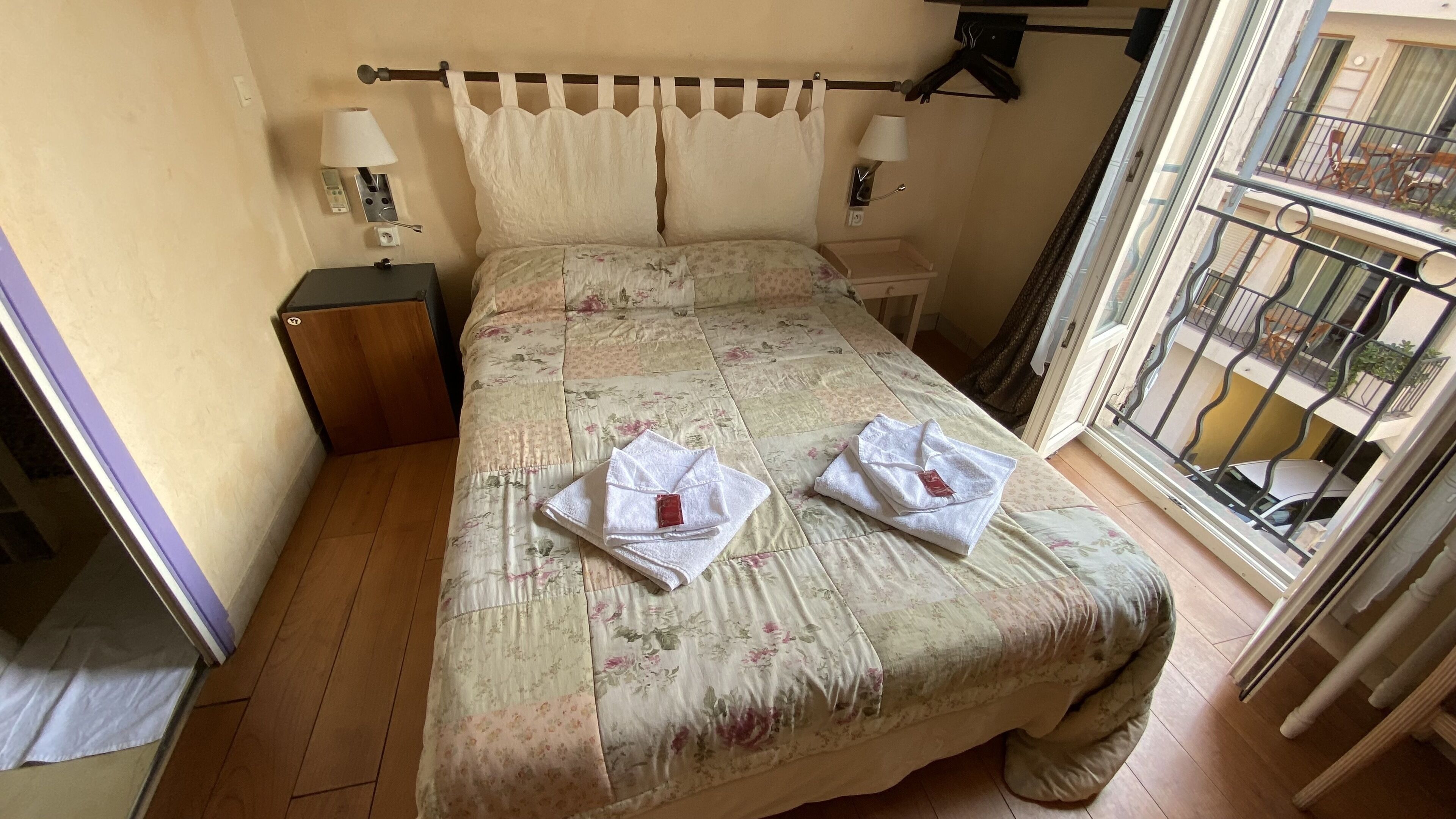 Un dormitorio con una colcha estampada de flores, dos lámparas de noche, una mesita de noche y vistas al balcón a través de una ventana.
