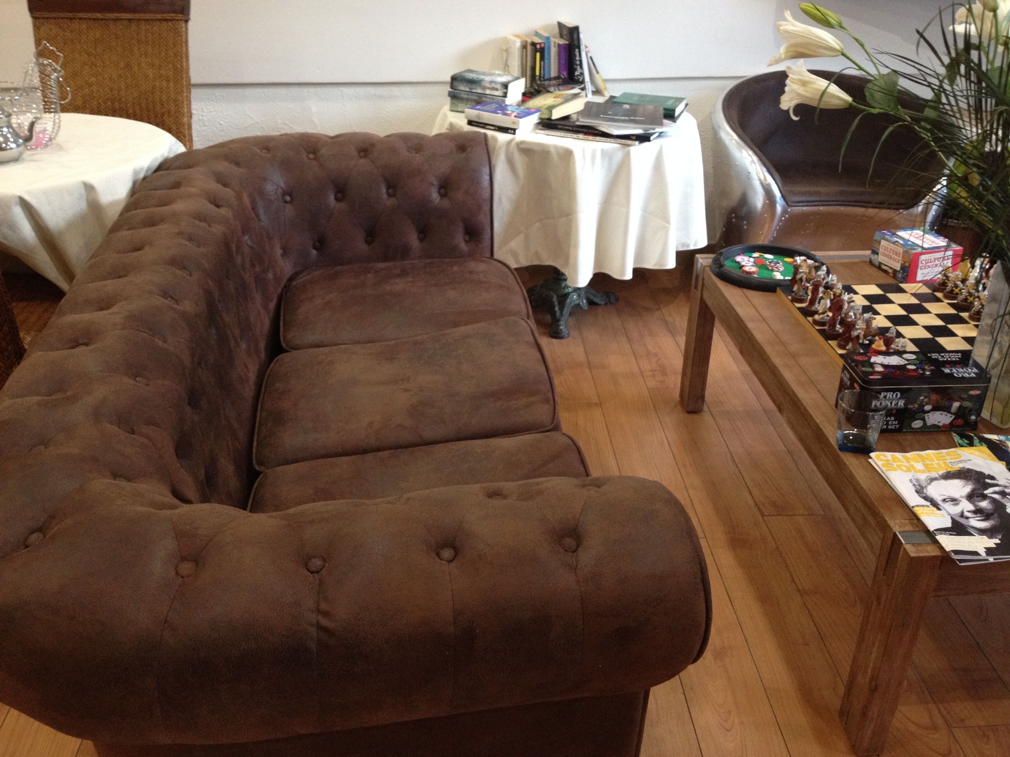 A brown tufted sofa in a room with wooden flooring, a table with books and a board game, and a vase with white flowers.
