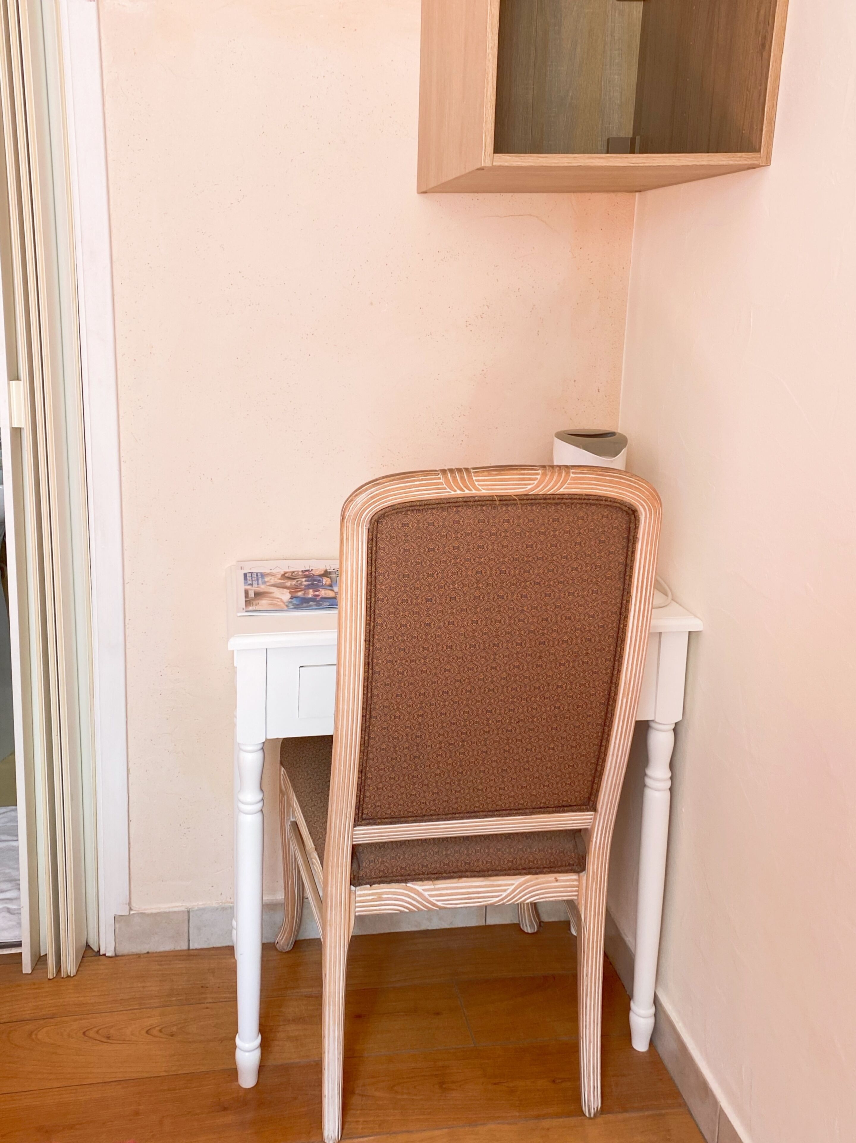 A corner of a room with a white chair with a patterned cushion, a small table with a book, and a wooden cabinet above.