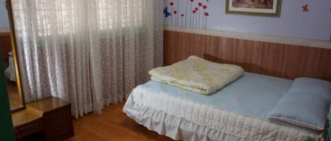 Un dormitorio con suelo de madera, una cama con colcha blanca y almohada azul, una mesita de noche de madera, un espejo y una ventana con cortinas translúcidas.