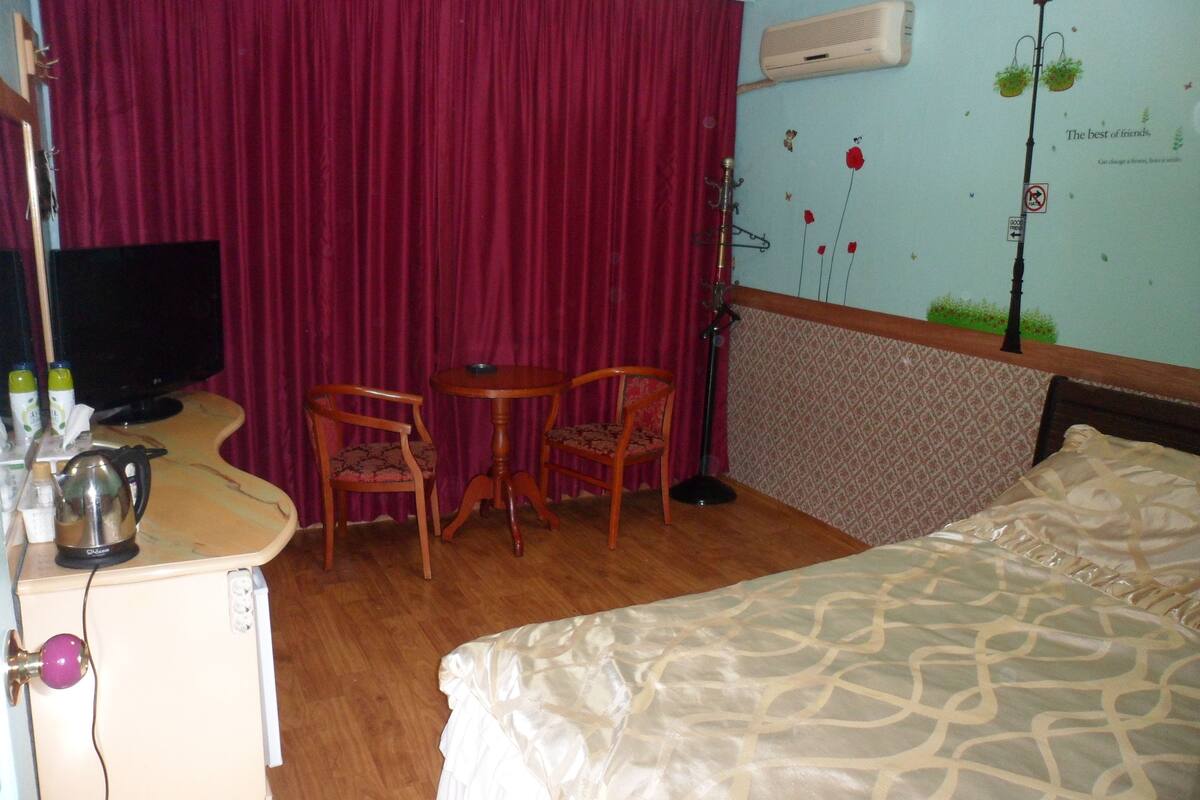 Une chambre d’hôtel avec un lit, un bureau avec un ordinateur, deux chaises, une petite table et un rideau rouge.