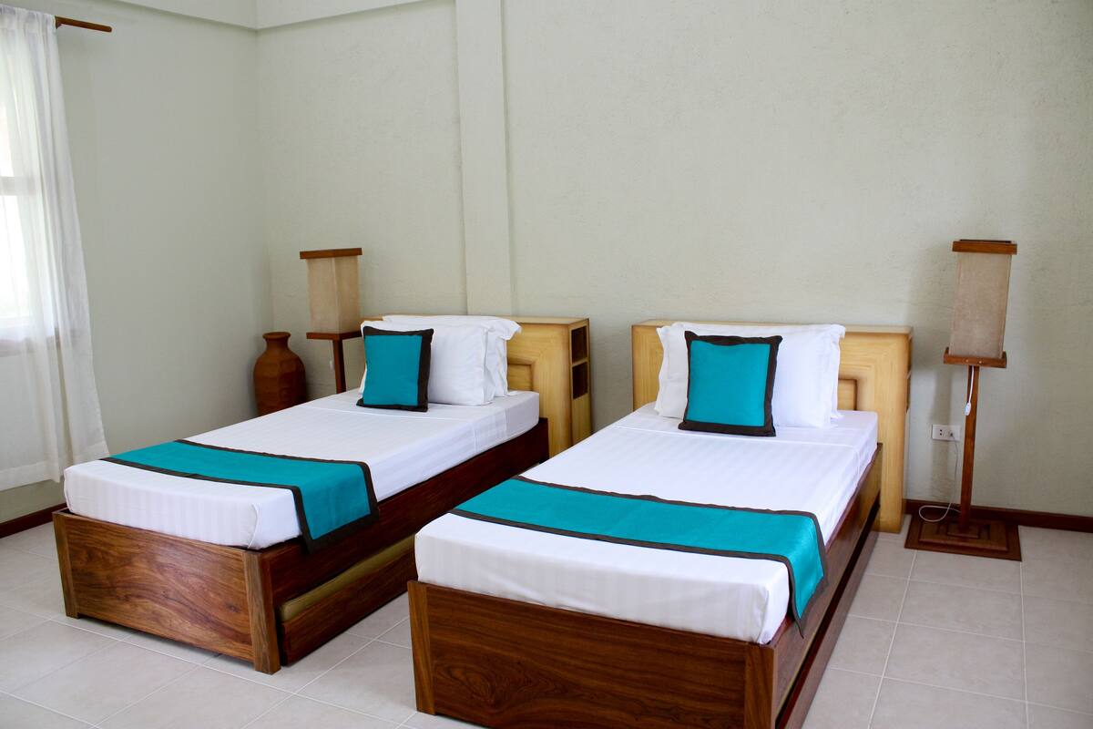 Deux lits en bois, recouverts de draps blancs et agrémentés de touches de couleur turquoise, une table de chevet en bois et une lampe sur pied dans une pièce aux murs blancs et au sol carrelé.