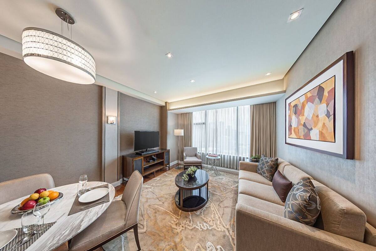 A modern hotel room with a dining area, a sofa, a television, and a large window.