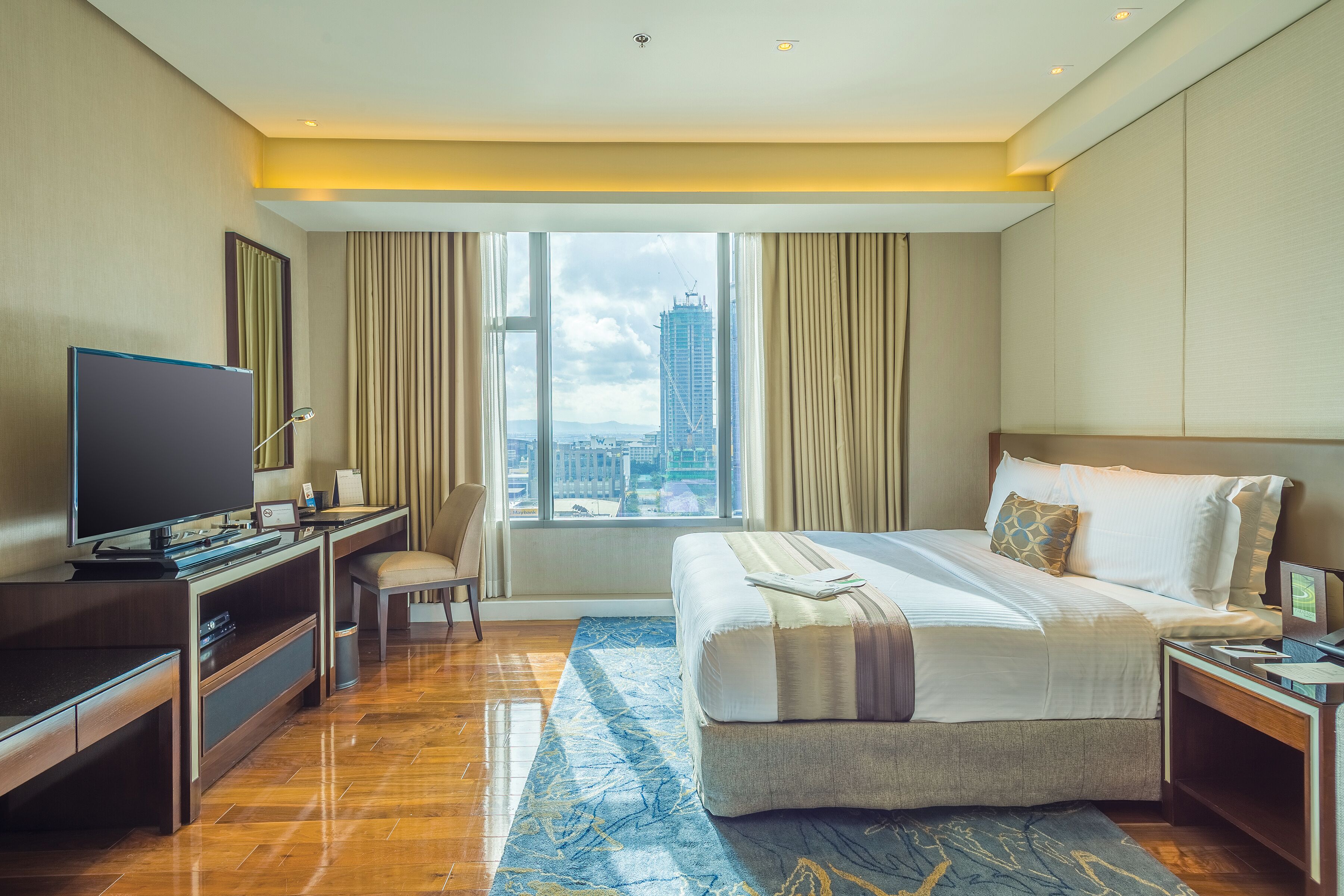 A hotel room with a large bed, a desk with a flat-screen TV, a chair, and a view of a cityscape through a large window.