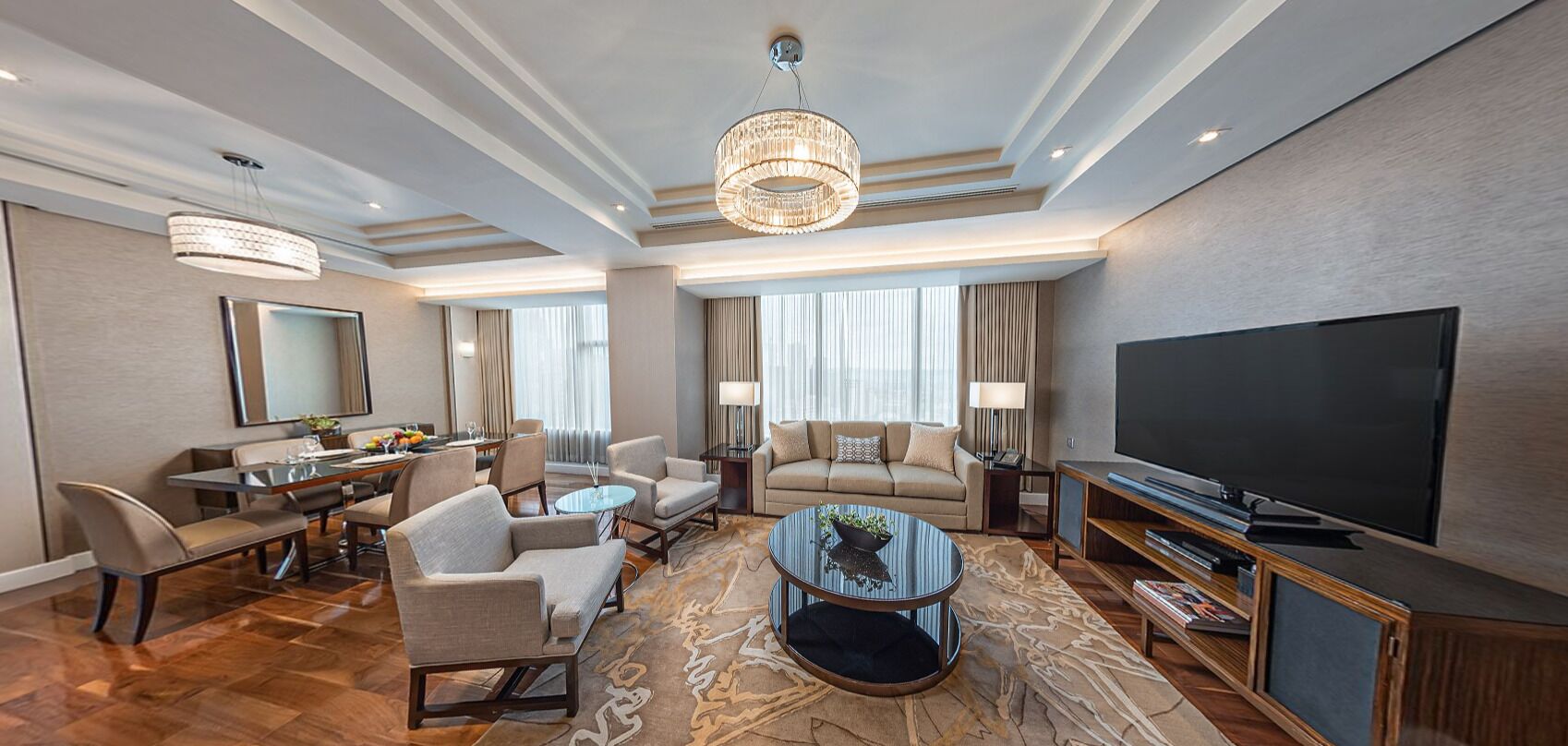A spacious hotel room with a large flat-screen TV, a dining area with a table and chairs, a sofa, a coffee table, and a chandelier.