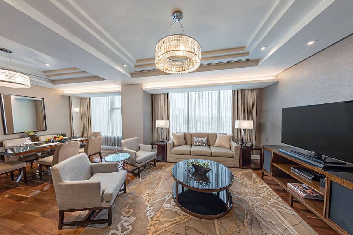A spacious hotel room with a large flat-screen TV, a dining area with a table and chairs, a sofa, a coffee table, and a chandelier.