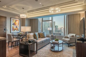 Uma sala de estar moderna com sofá, duas poltronas, mesa de centro e TV. Há grandes janelas com vista para a cidade, um candelabro e um quadro abstrato emoldurado na parede.