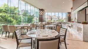 Un restaurante espacioso con techos altos y ventanas amplias que ofrecen vistas a la ciudad. Las mesas están dispuestas con manteles blancos y sillas.