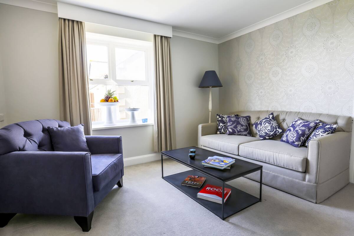 A living room with a dark blue sofa, a light beige sofa with patterned cushions, a coffee table with books, a floor lamp, and a window with curtains.