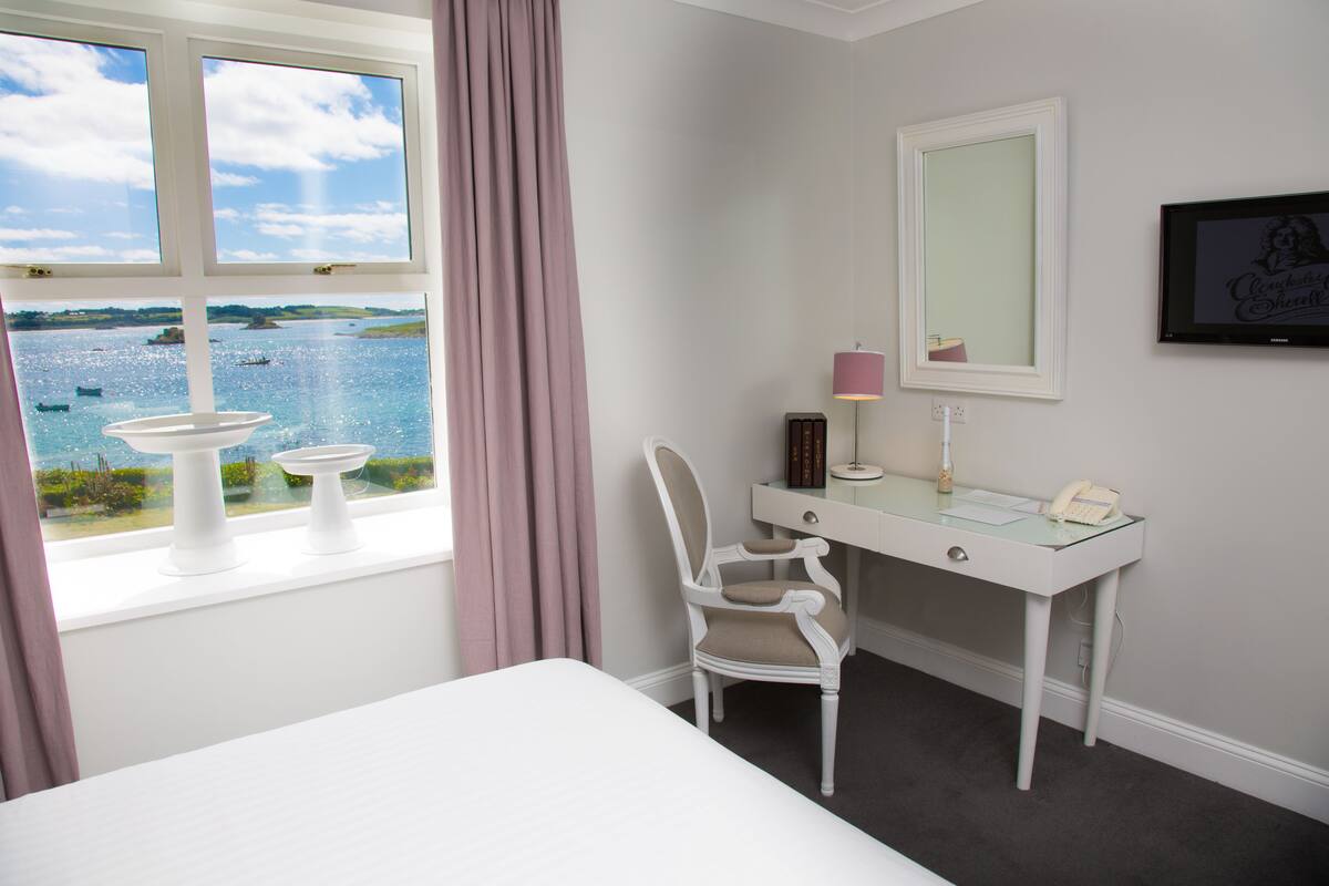 A bedroom with a bed, a desk with a chair, a mirror, and a view of the sea through a window.