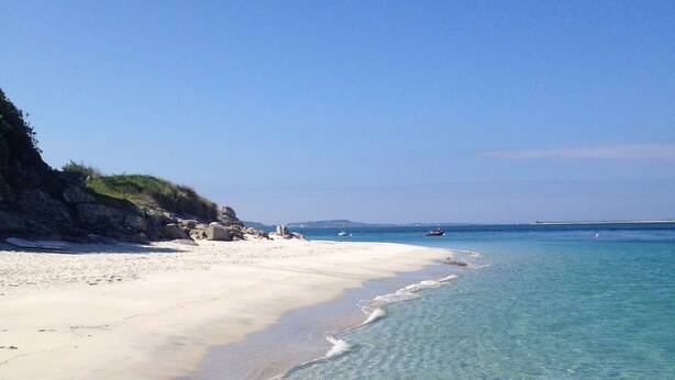 Una playa con aguas transparentes y poco profundas, una orilla arenosa y afloramientos rocosos.