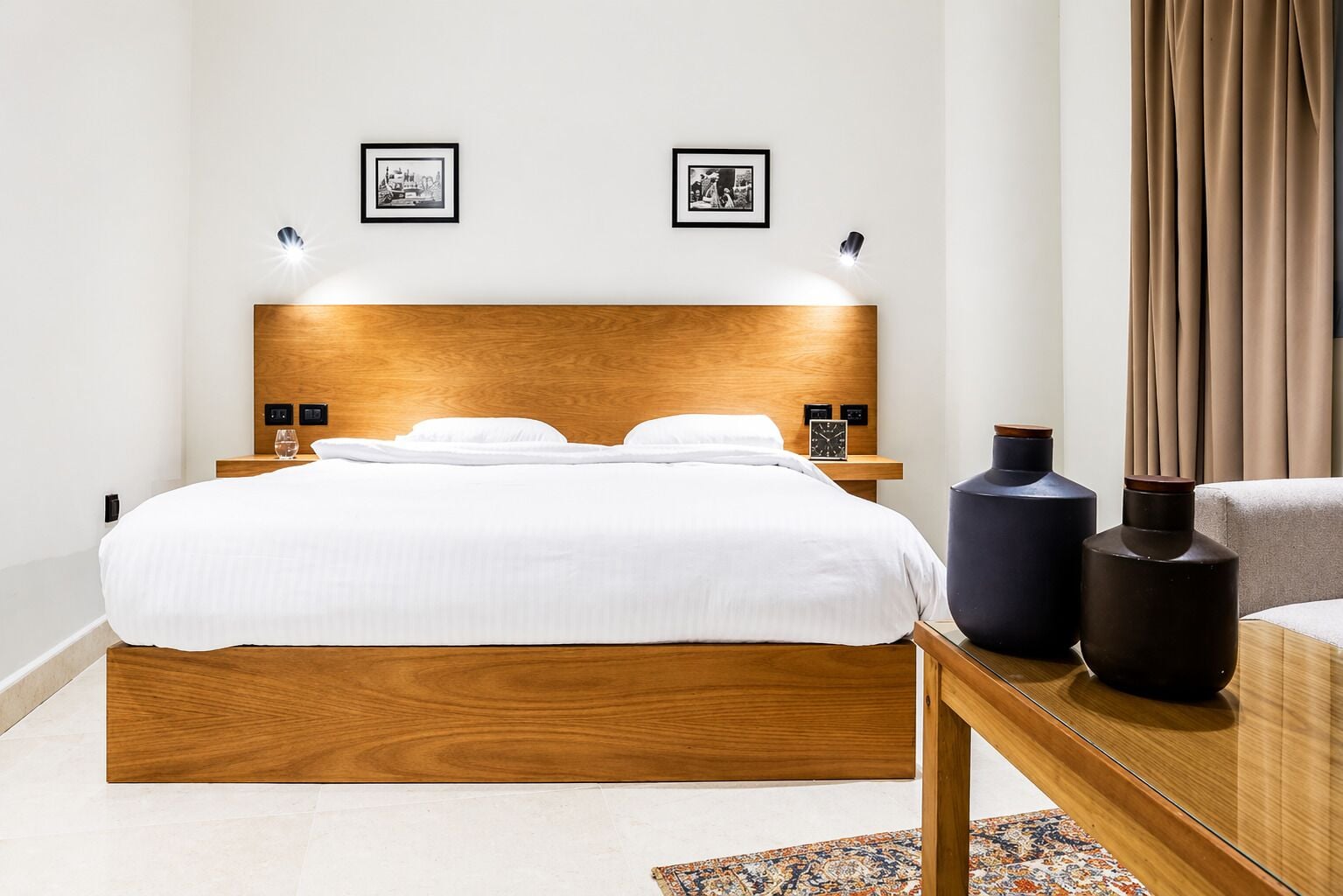 A modern hotel room with a wooden headboard, a large bed, bedside tables with lamps, two framed pictures on the wall, and a nightstand with a clock.