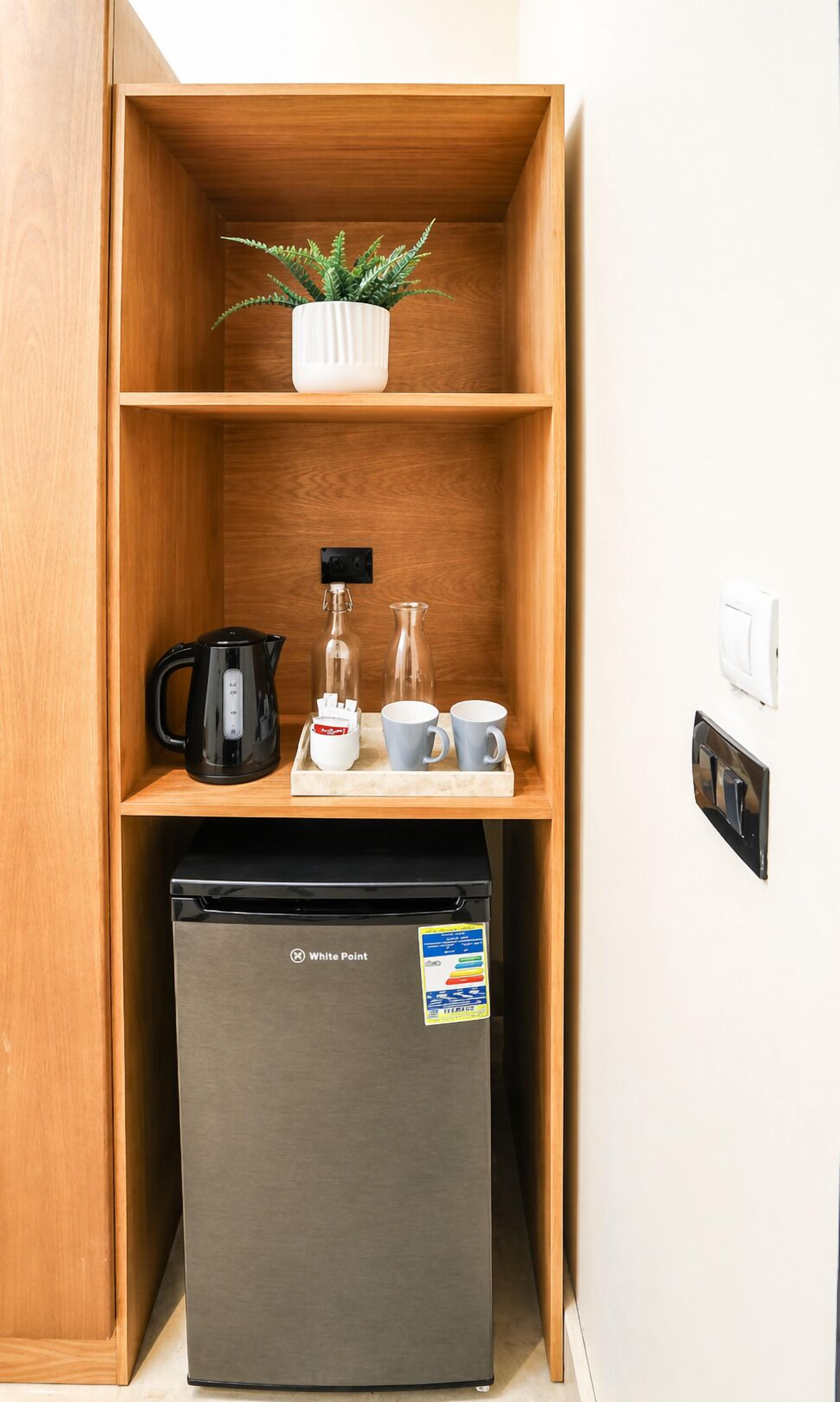 Una kitchenette compacta con un refrigerador, una cafetera, un hervidor de agua, una botella de vidrio y una planta en maceta.