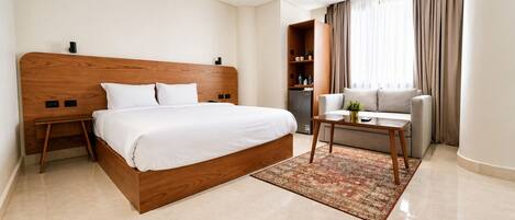 A hotel room with a wooden bed, a small table, a sofa, a bookshelf, and a large window with curtains.