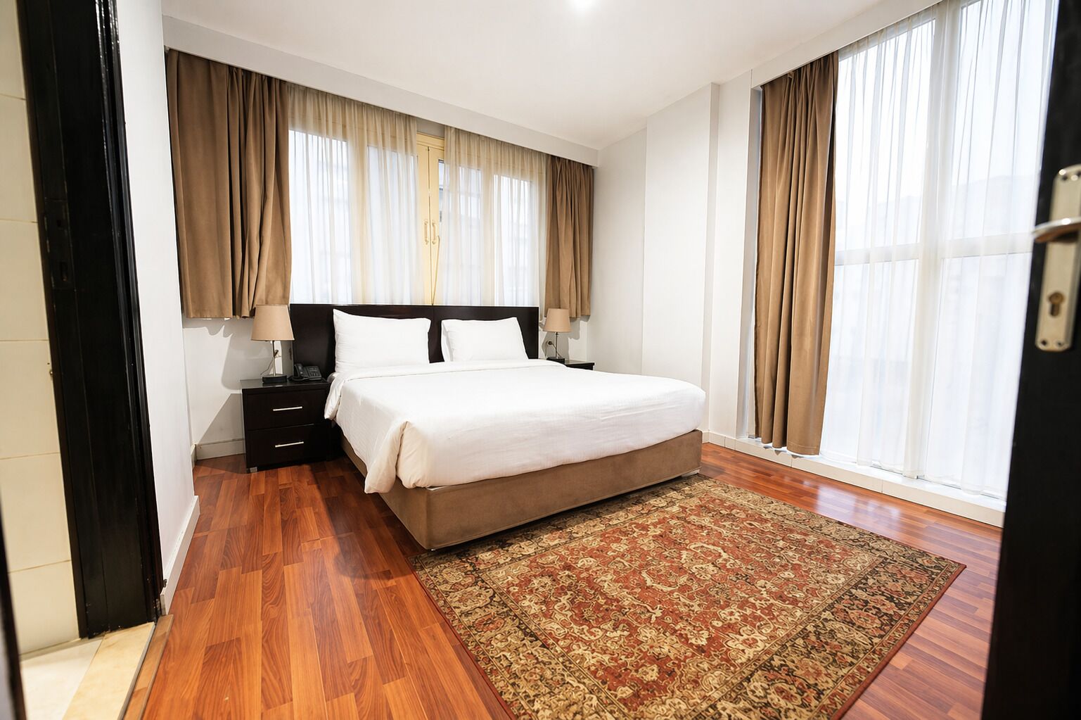 A hotel room with a large bed, a nightstand, a desk, and a window with curtains.