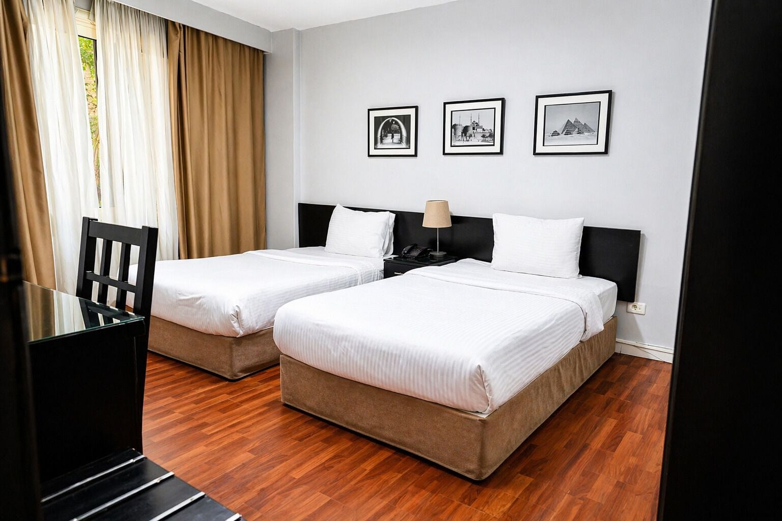 A hotel room with two beds, a desk, and a chair. There are three framed pictures on the wall.