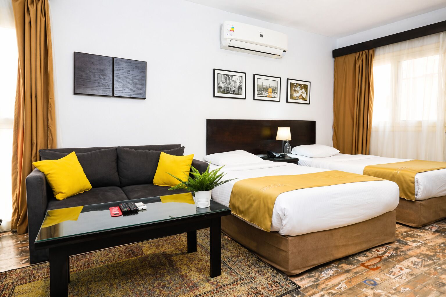 A hotel room with a sofa, two beds, a coffee table, a plant, and artwork on the wall.