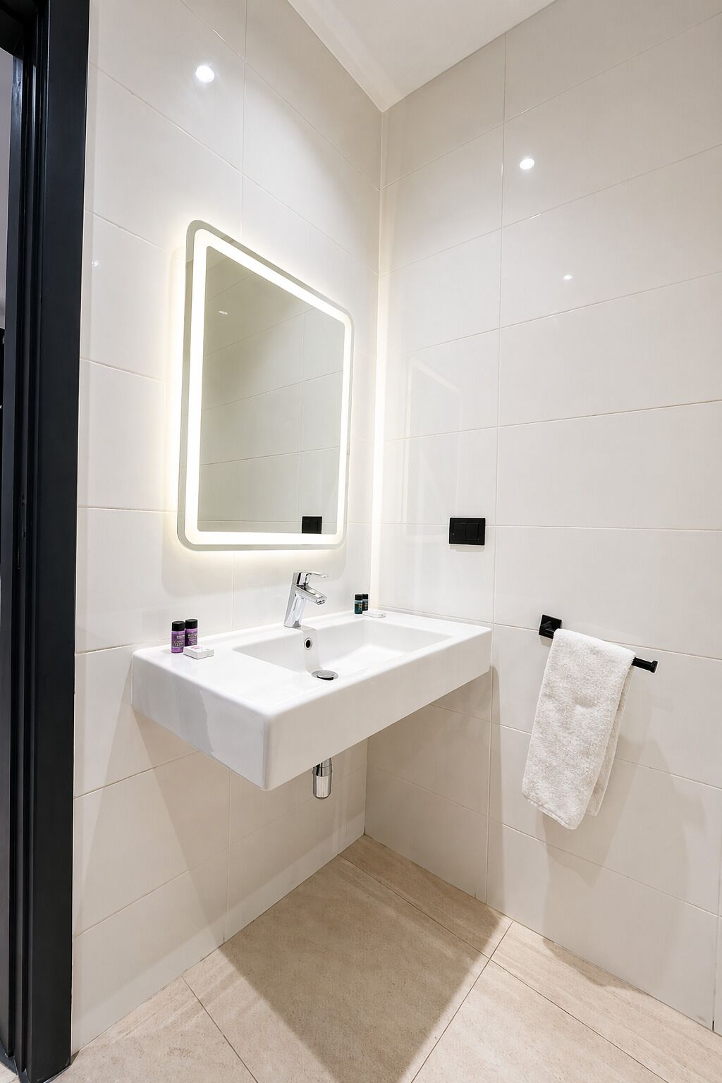 Un baño moderno con un lavamanos blanco, un espejo con bordes iluminados y un perchero para toallas.