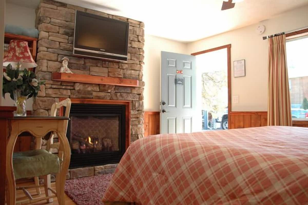 A bedroom with a stone fireplace, a television mounted above it, a bed with a plaid bedspread, and a door leading outside.