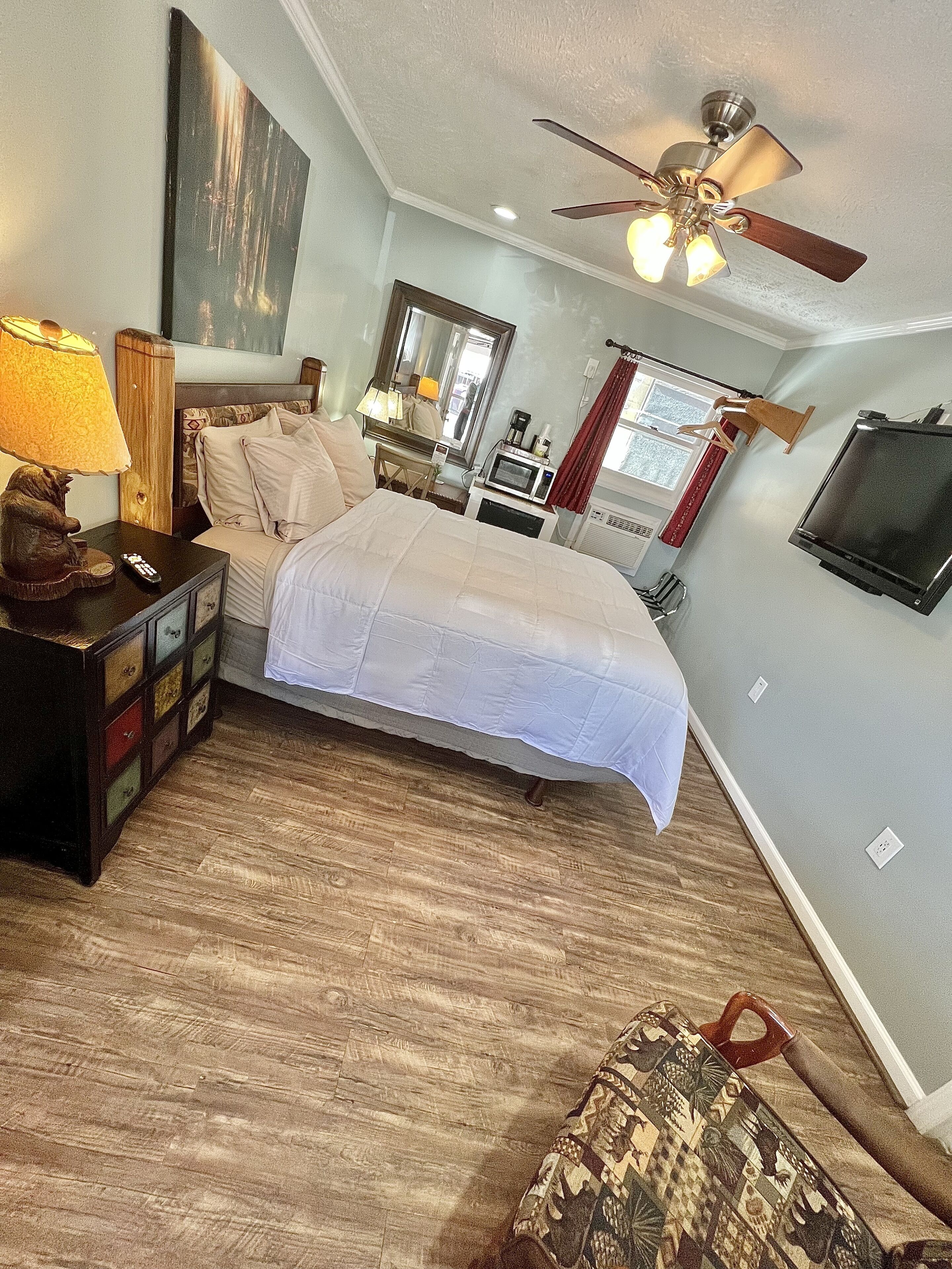 A bedroom with a bed, a nightstand, a lamp, a dresser, a ceiling fan, a TV, and a window with curtains.