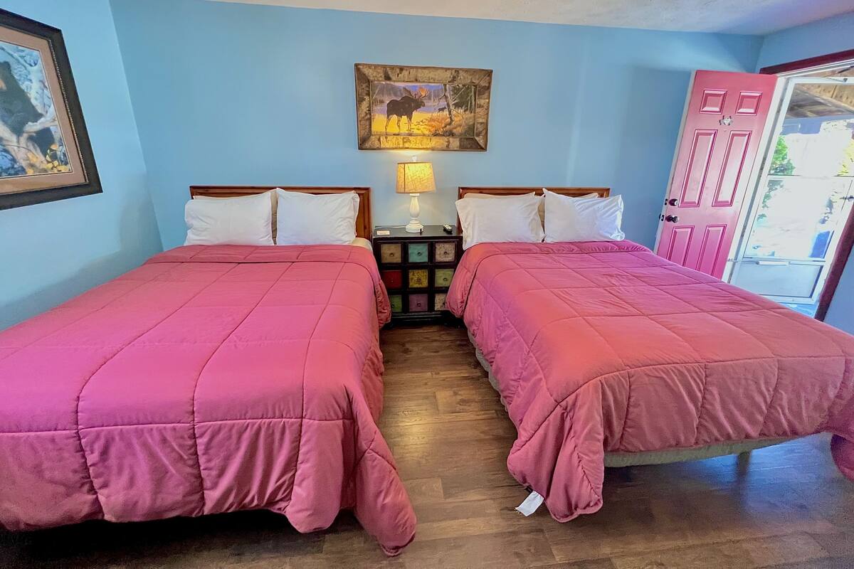 A room with two beds, a nightstand, a lamp, a framed picture, and a door.