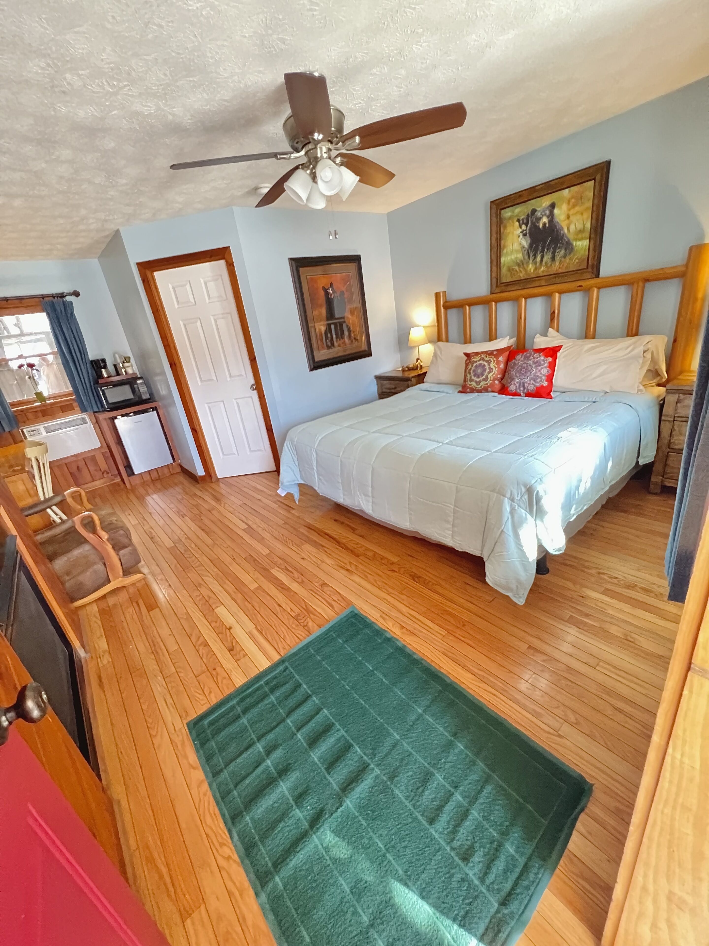 A bedroom with a bed, wooden floor, ceiling fan, and a green rug.