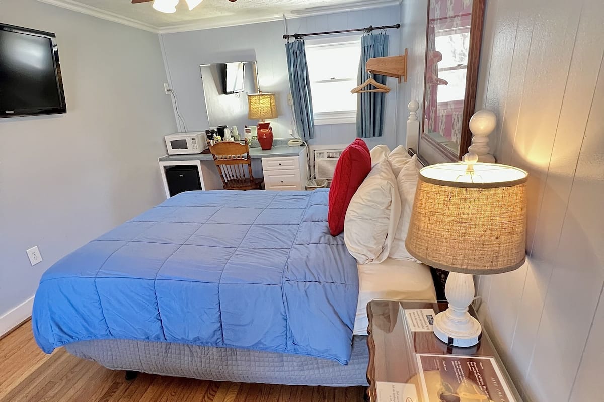 A bedroom with a bed, a ceiling fan, a bedside table with a lamp, a dresser, a desk with a chair, a TV, and a window with curtains.