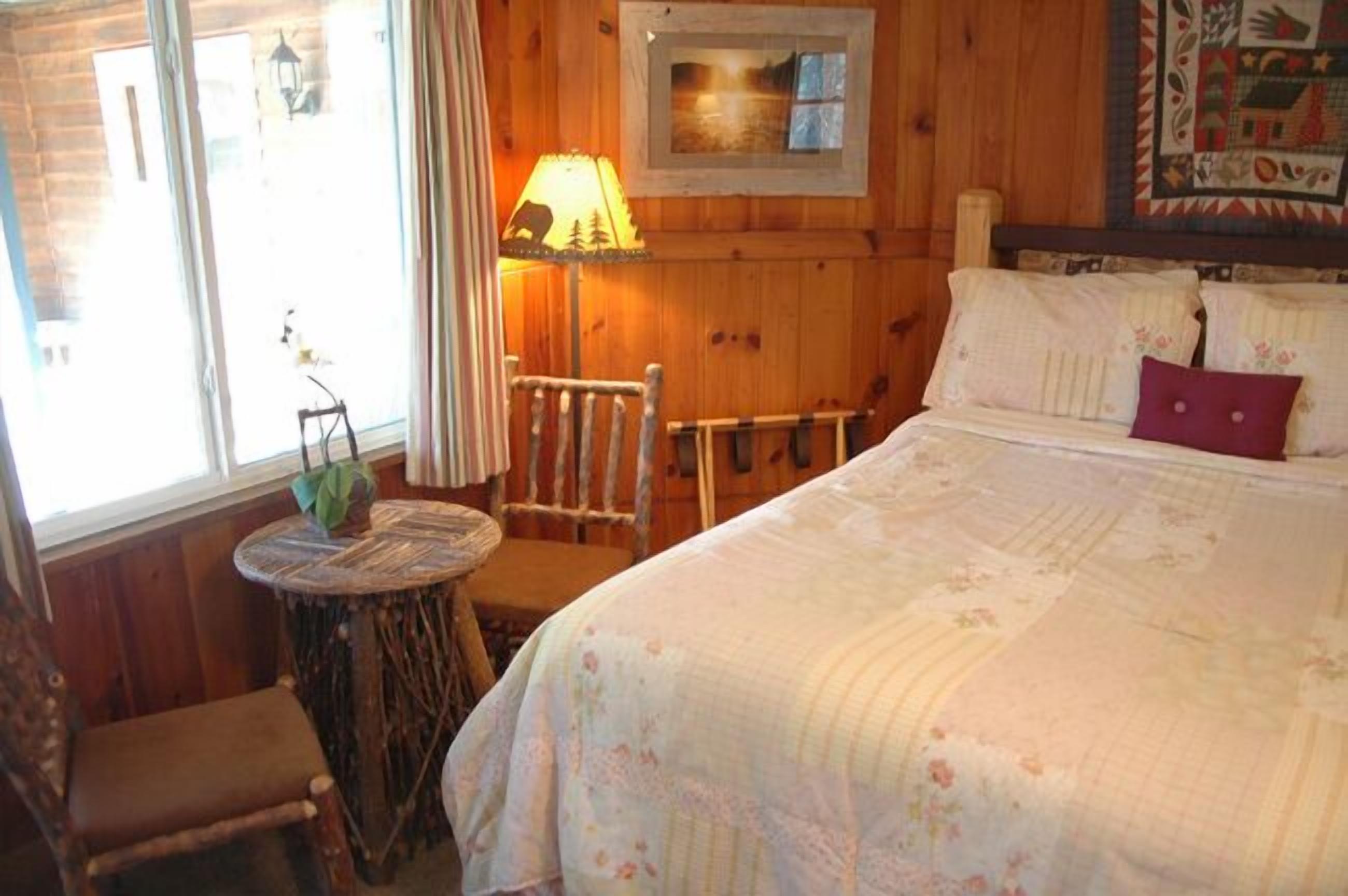 Een knus ingerichte kamer met een bed, een stoel, een klein tafeltje met een plant en een raam met gordijnen.