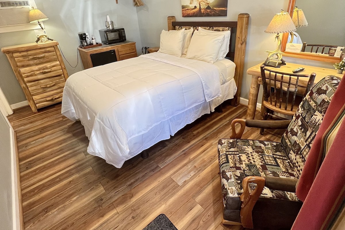 A bedroom with a bed, wooden floor, ceiling fan, bedside table, lamp, and a chair.