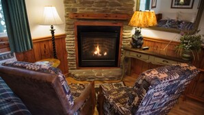 A cozy living room with a stone fireplace, two patterned armchairs, a wooden coffee table, and a lamp.