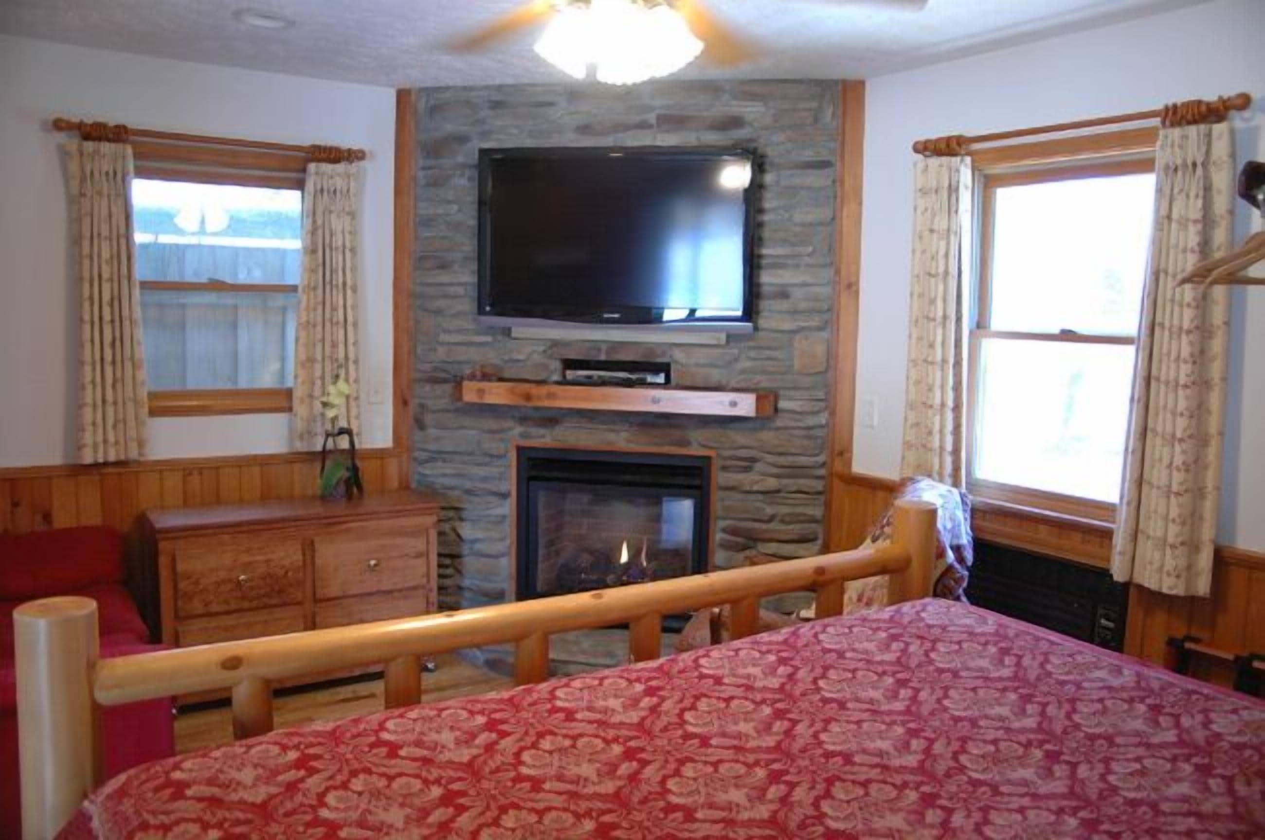 A bedroom with a stone fireplace, a television mounted above it, a wooden bed with a red patterned bedspread, and a wooden dresser.