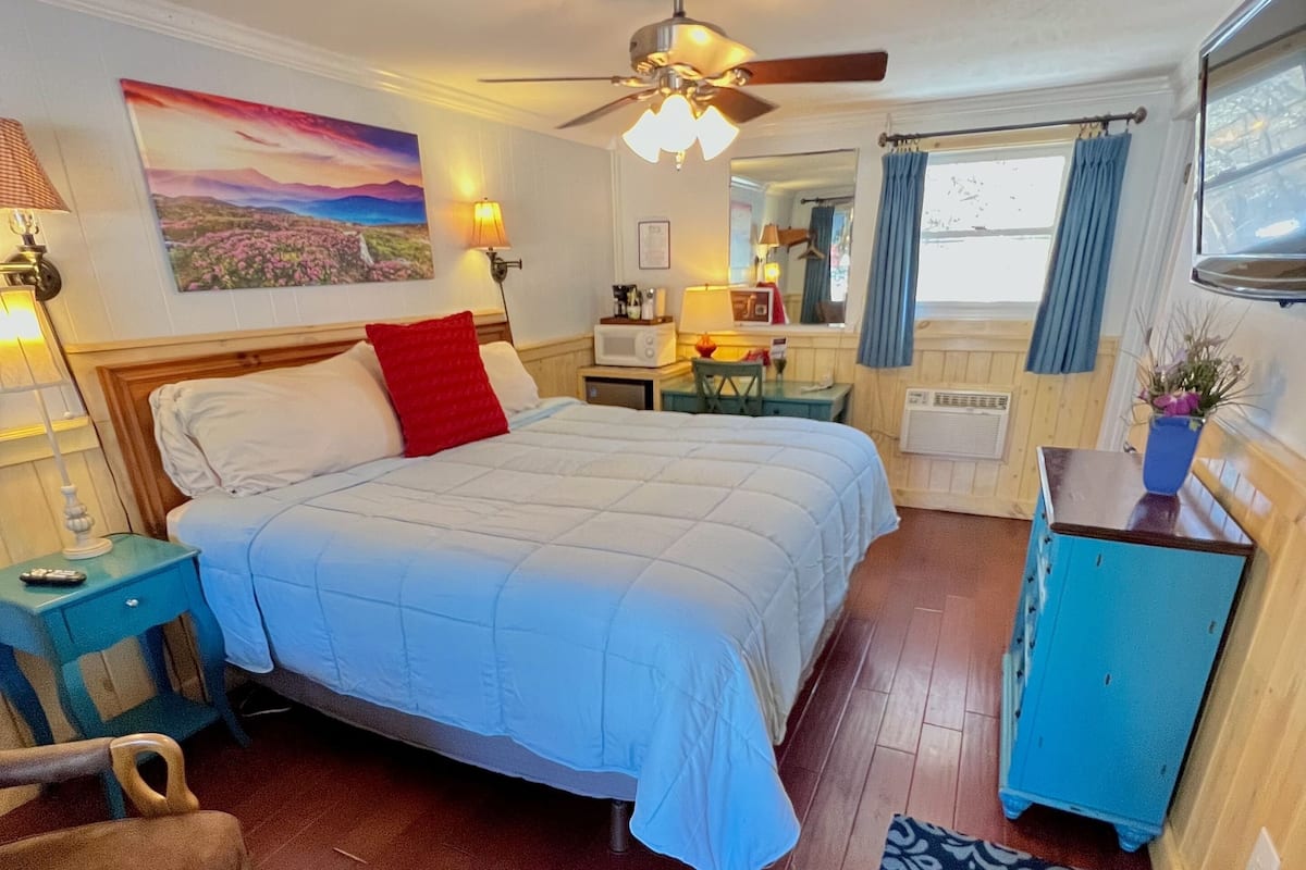 A bedroom with a bed, a desk, a chair, a ceiling fan, a window with curtains, and a wall-mounted mirror.