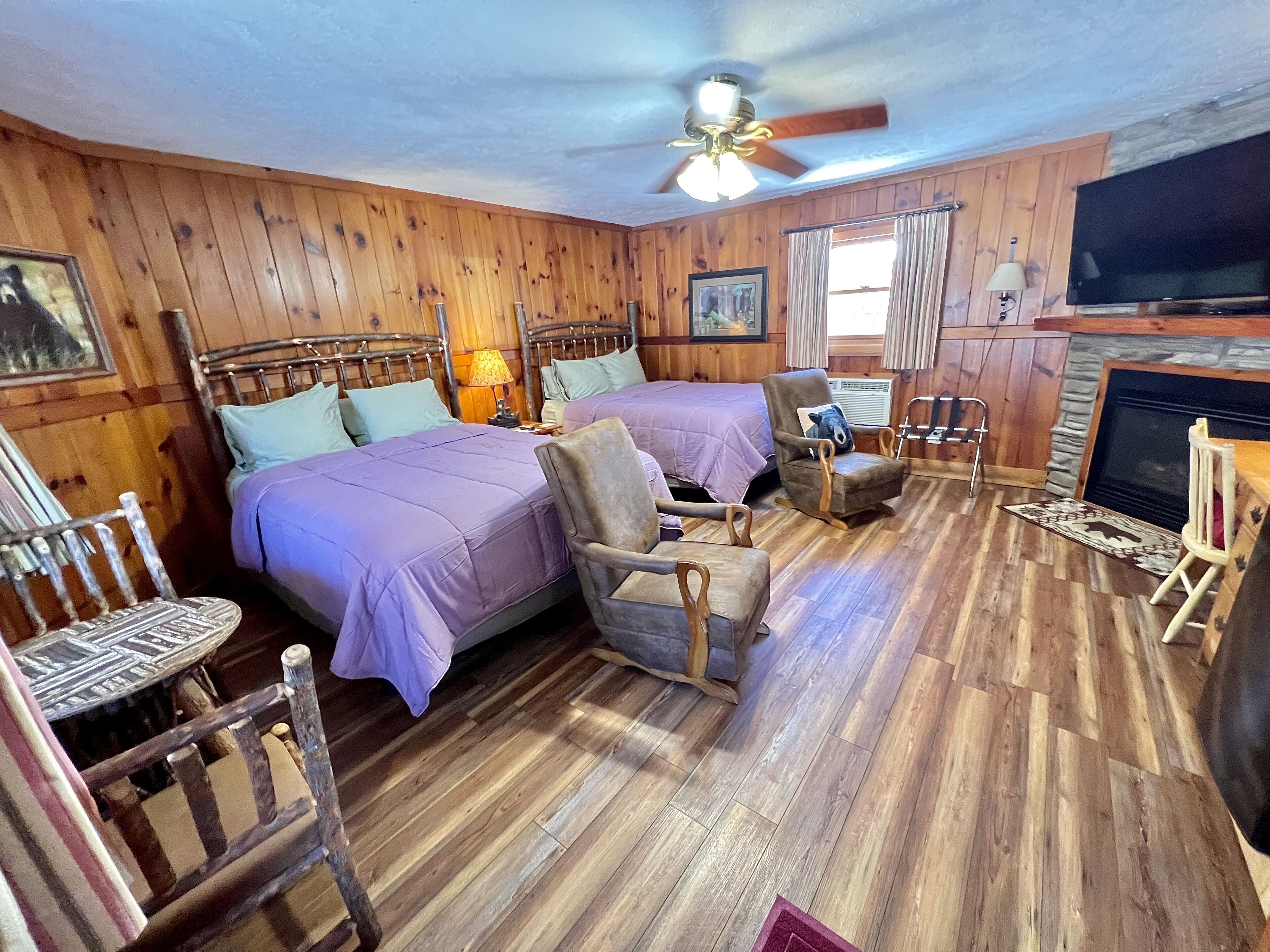A cabin-style bedroom with two beds, a wooden floor, a ceiling fan, a fireplace, and a television.
