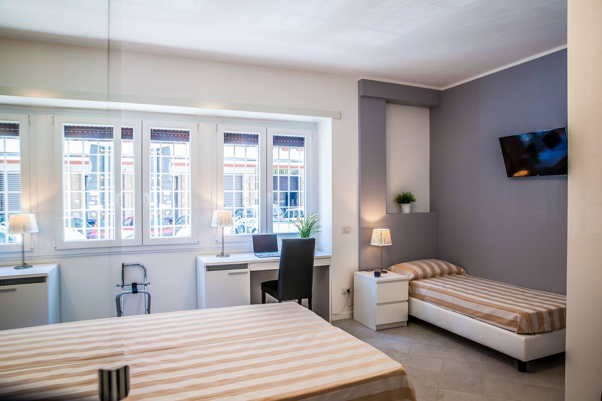 A room with a bed, a desk with a computer, a chair, a nightstand, and a TV.