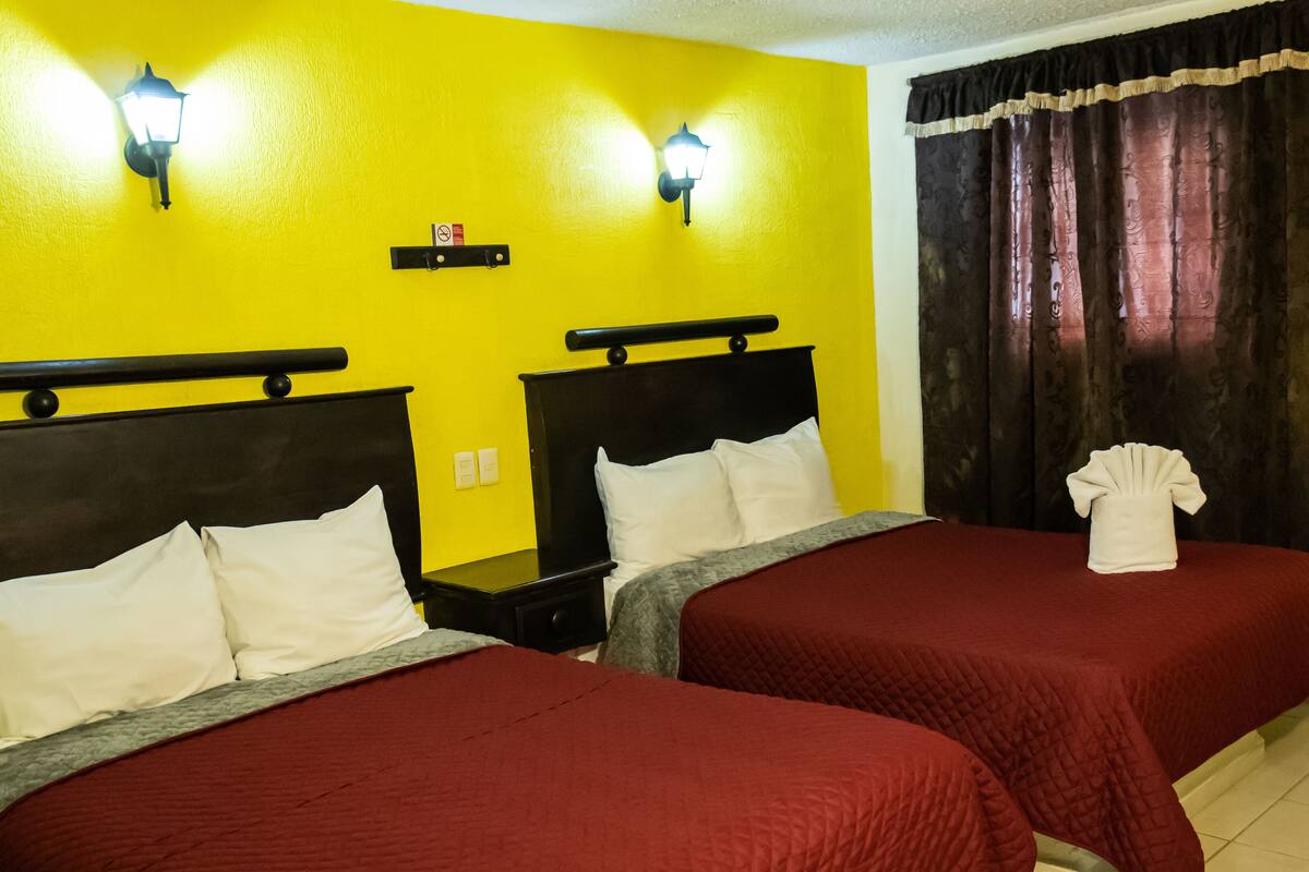 A hotel room with two beds, each with a red bedspread and white pillows, against a yellow wall with two wall-mounted lights.