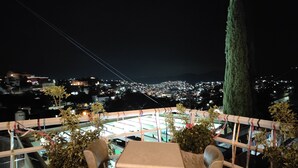 Terraza en la azotea con mesa y sillas, rodeada de plantas en macetas, con vistas a un paisaje urbano nocturno.