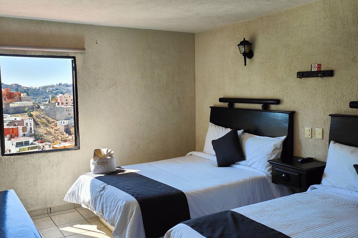 A hotel room with two beds, a window with a city view, a desk, and a nightstand with a lamp.