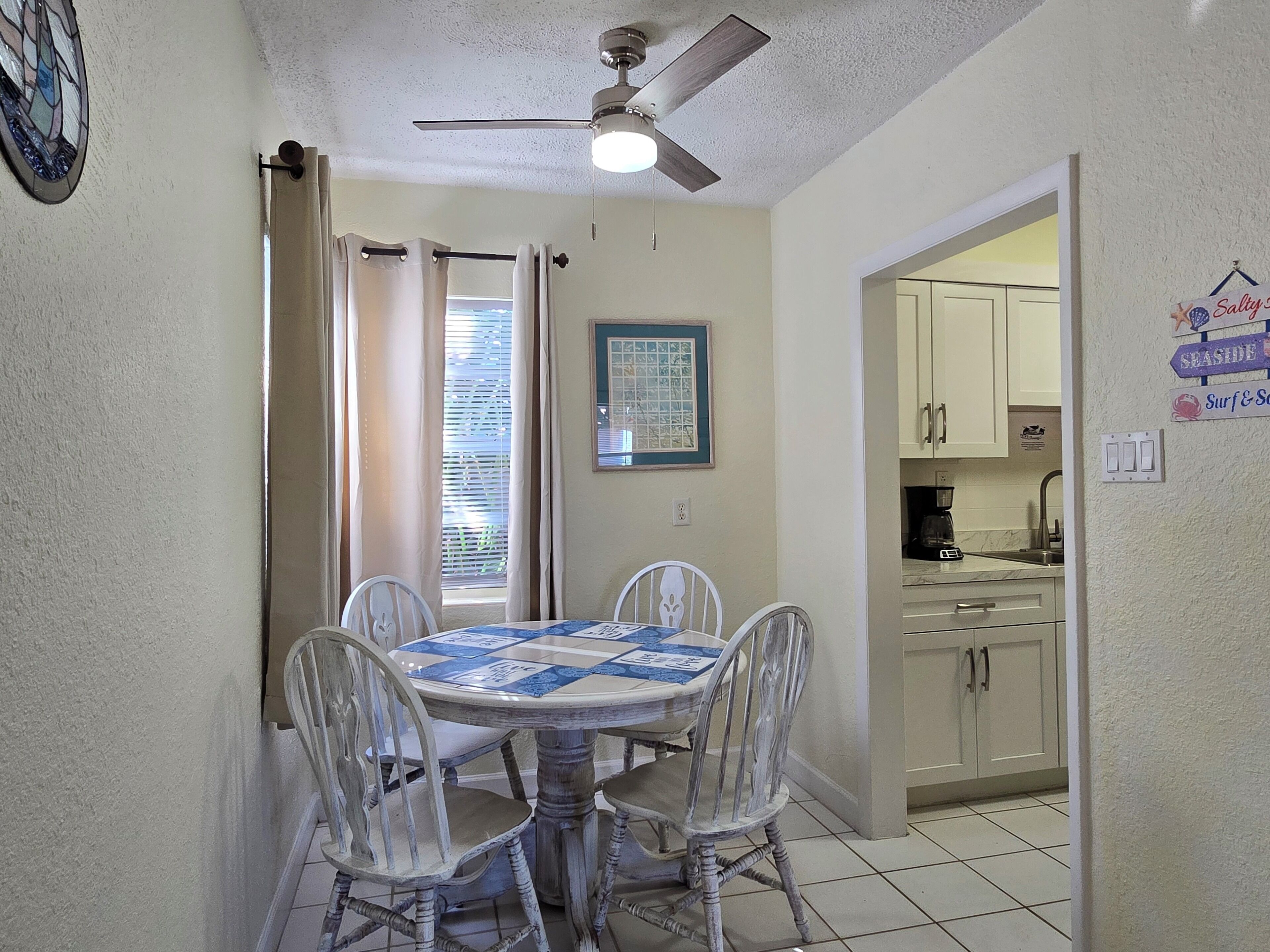 A dining area with a round table and four chairs, a ceiling fan, a window with curtains, and a kitchen visible through an open door.