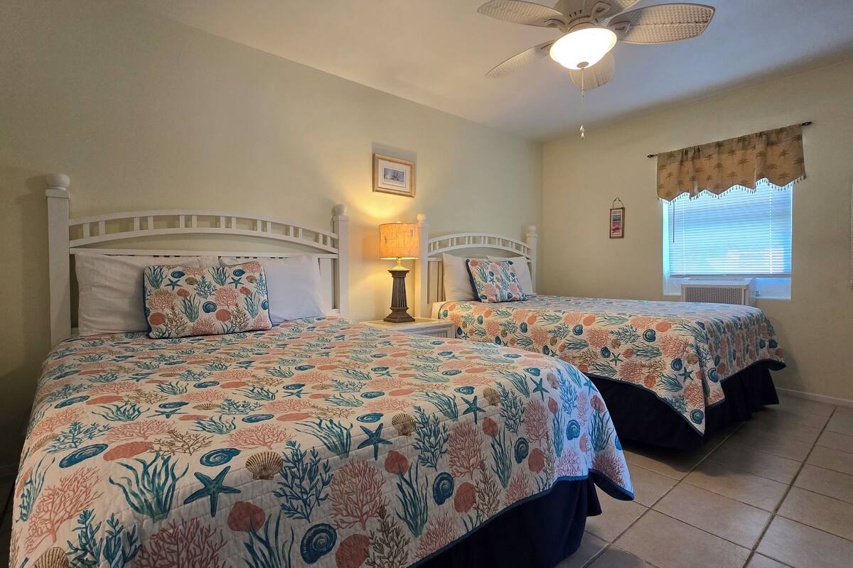 Habitación de hotel con dos camas, cada una con cubrecama estampado con motivos florales, un ventilador de techo, una lámpara de noche y una ventana con persianas.
