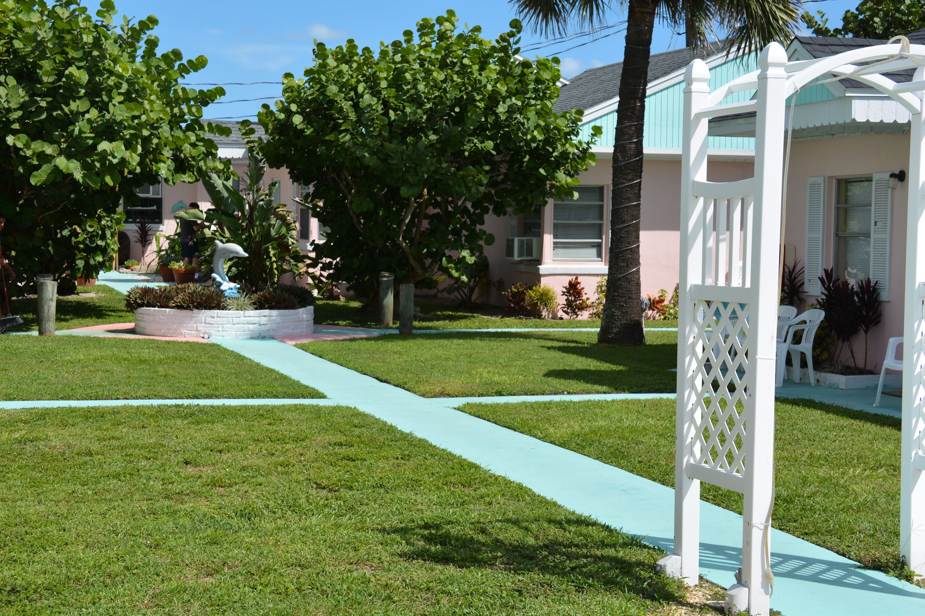 Un jardín cuidado con un sendero, una pérgola blanca y un edificio con fachada azul claro.