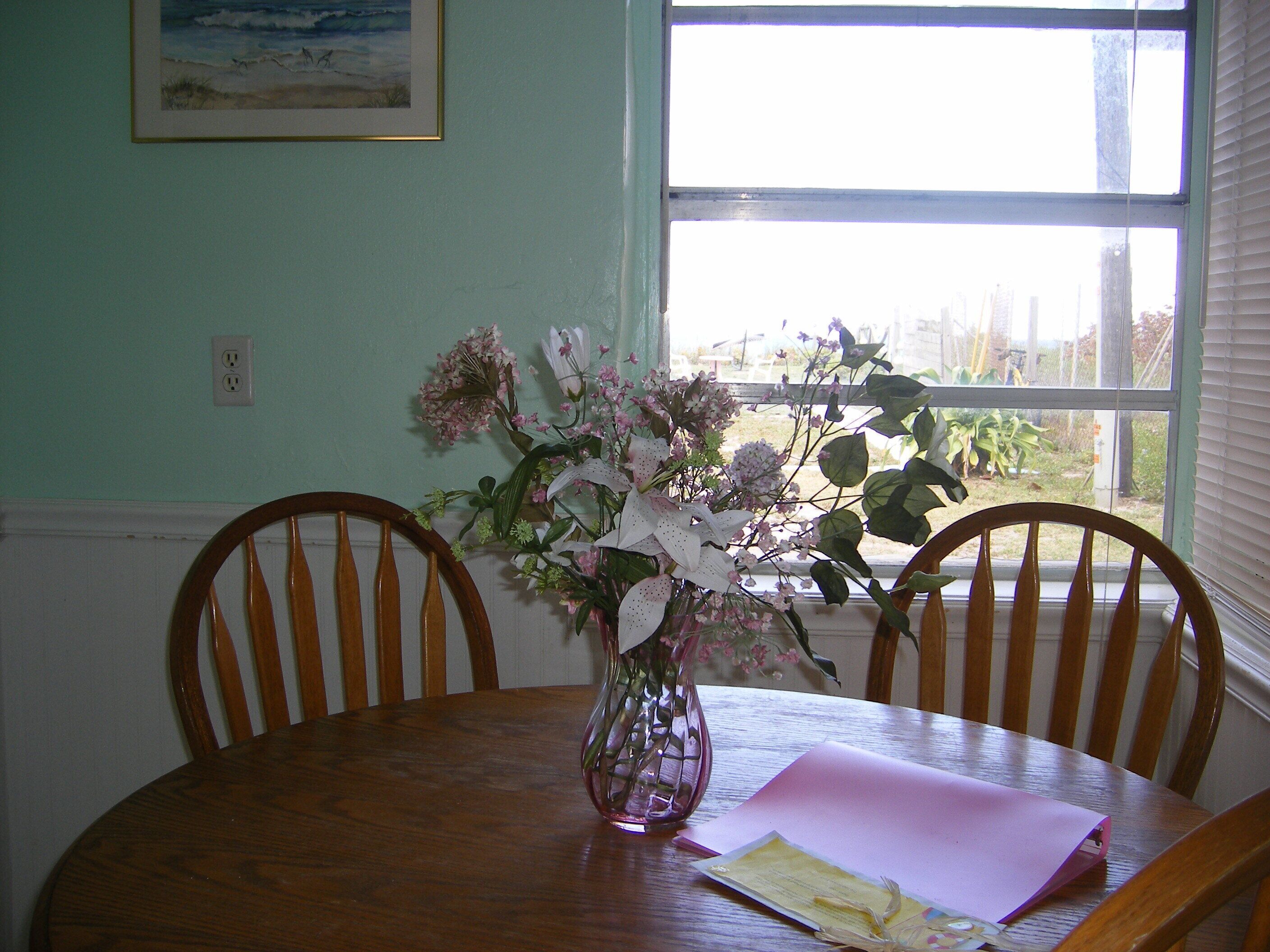 A dining table with a vase of flowers, a pink envelope, and a framed picture on the wall.