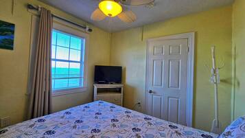Un dormitorio con cama, ventilador de techo, televisor sobre un soporte, puerta y ventana con cortinas.