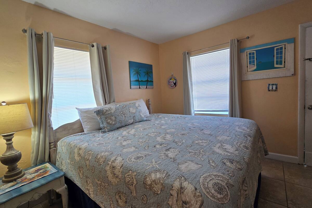 Un dormitorio con cama, cortinas, un cuadro y una lámpara.