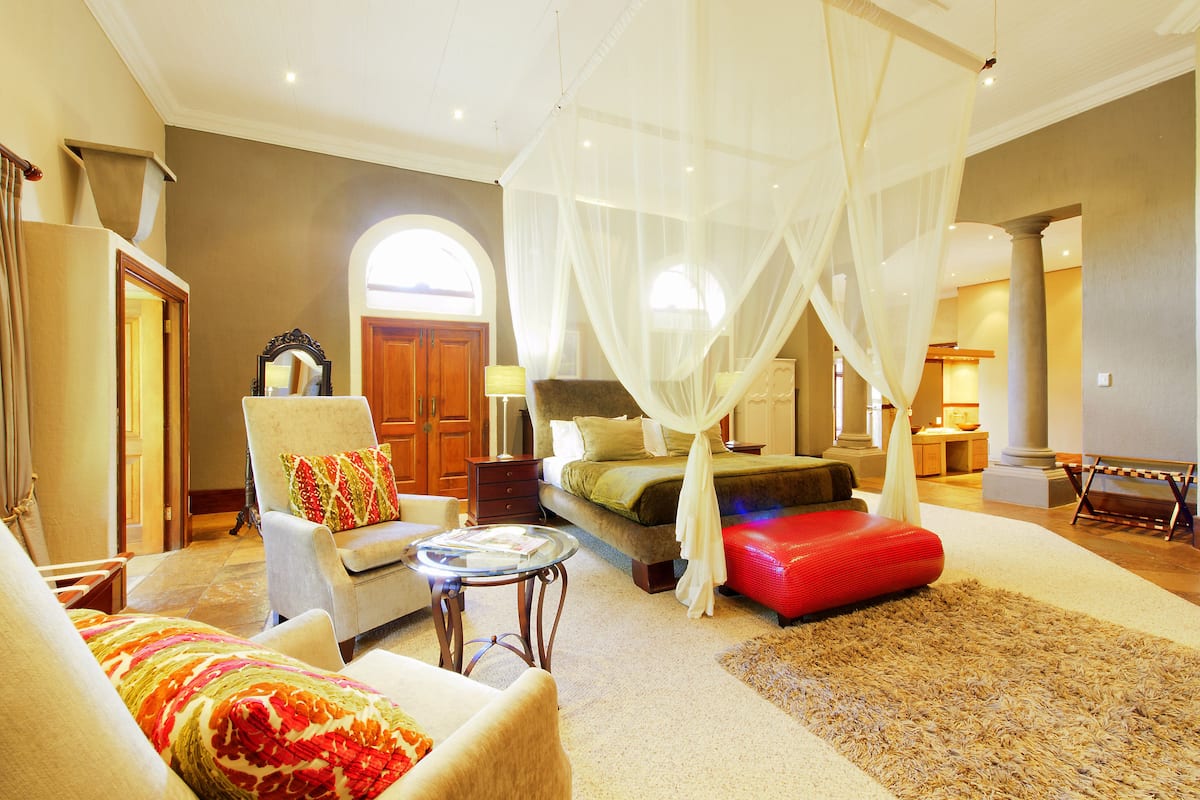 A spacious bedroom with a canopy bed, a sitting area with a chair and a small table, and a large arched doorway.