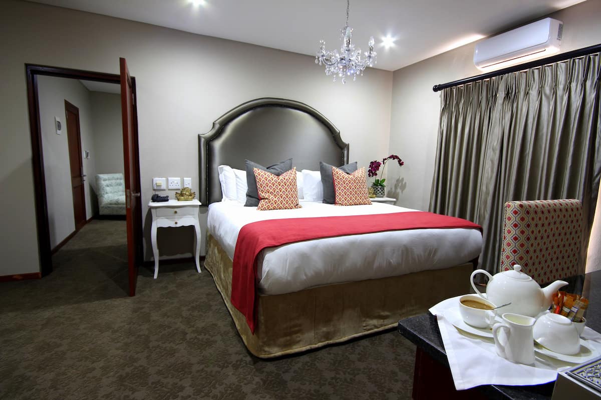 A hotel room with a large bed, a bedside table with a lamp, a chair, and a table with a teapot and cups.